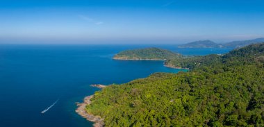 Tayland 'ın Phuket kentindeki tropikal kıyı şeridinin hava manzarası, yoğun yeşillik, canlı mavi bir deniz, hareketli bir tekne ve uzak orman tepeleri..