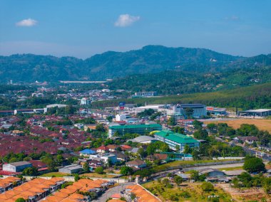 Phuket, Tayland 'da kırmızı kiremitli çatılar, yeşil tepeler, modern binalar ve arka planda açık mavi bir gökyüzü bulunan bir kasabanın hava manzarası..
