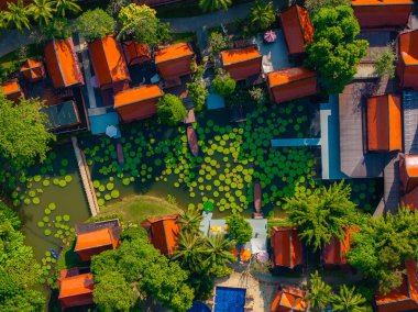 Büyük yapraklı bir nilüfer havuzu manzarası, ahşap geçitler, ve yemyeşil yeşilliklerle çevrili kırmızı turuncu çatılı Tayland tarzı binalar..