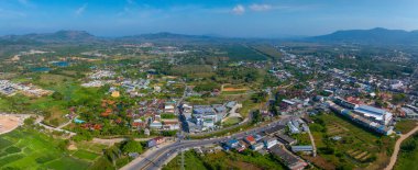 A vibrant aerial view of a town in Phuket, Thailand, featuring buildings, roads, fields, and distant hills under a clear blue sky. clipart