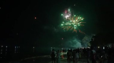Tayland 'da gece gösterişli bir plaj sahnesi, kalabalık, parlayan fenerler, havai fişekler, parıldayan su yansımaları, ve ağaç siluetleri..