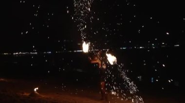 Bir sanatçı, gece sahilin birinde, suya yansıyan kıvılcımlar ve ışık ile yangın aksesuarlarını manipüle ederek neşeli ve dinamik bir sahne yaratır..