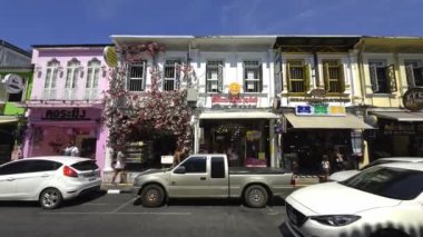 Phuket Old Town, Tayland 'da canlı bir cadde, renkli dükkanlar, pembe bir kafe, dükkanlar, park edilmiş arabalar ve parlak mavi gökyüzü altında yayalar..