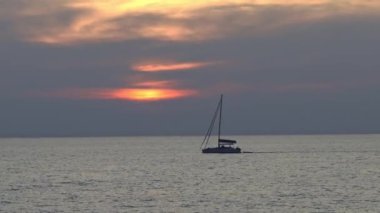Tayland, Phuket yakınlarındaki sakin okyanus suları üzerinde sakin bir günbatımı, gökyüzünde canlı turuncu ve sarı renkte bir yelkenli..