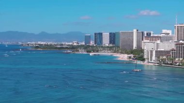 Honolulu, Hawaii 'deki Waikiki Sahili' nin havadan görünüşü turkuaz sular, kumlu sahil şeridi, yüksek oteller ve okyanusta süzülen bir yelkenli gösteriyor..