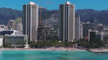 Honolulu, Hawaii 'deki Waikiki Sahili' nin havadan görünüşü turkuaz suları, sahile gidenleri, yüksek binaları ve arka planda yemyeşil dağları gösteriyor..