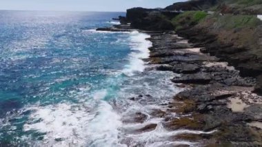 Oahu 'nun kayalık kıyı şeridinin havadan görüntüsü. Volkanik kayalara çarpan dalgalar, turkuaz sular ve dolambaçlı bir sahil yolunda seyahat eden araçlar..
