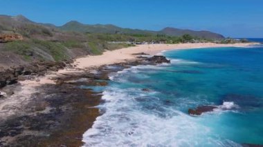 Hawaii, Oahu adasındaki bir kumsal manzarası, turkuaz sular, kayalık çıkıntılar, yumuşak dalgalar, yemyeşil yemyeşil ve uzak dağlar..