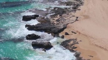 Dinamik dalgalar Oahu 'nun engebeli kıyı şeridi boyunca uzanan karanlık volkanik kayalara çarpar, küçük kumlu bölgeler ve canlı turkuaz okyanus renkleriyle..