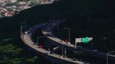 Oahu, Hawaii 'de bir otobanın hava görüntüsü, arabalar kıvrımlı bir üst geçitte ilerliyor, yemyeşil ve yoğun bir yerleşim alanı ile çevrili..