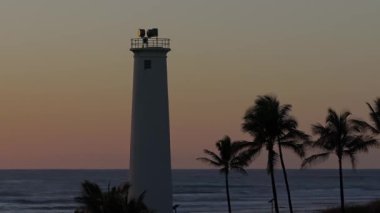 Oahu Adası, Hawaii 'de sakin bir günbatımı. Beyaz bir deniz feneri, sakin bir okyanus, eğimli bir gökyüzü ve ön planda yavaşça sallanan palmiye ağaçları..