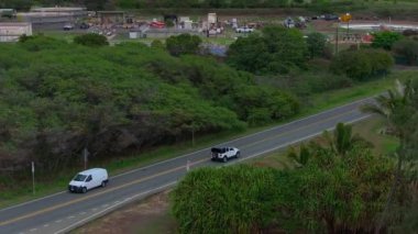 Oahu, Hawaii 'nin manzaralı manzarası, dolambaçlı bir yol, park edilmiş araçlar, yemyeşil bitki örtüsü, kayalık kıyı şeridi ve hareket halindeki nazik okyanus dalgalarını gösteriyor..