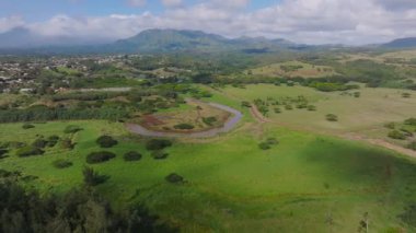 Hawaii 'deki Kauai adasının hava manzarası dolambaçlı bir nehir, yeşil tarlalar, yoğun bitki örtüsü, dağlar ve bulutlu gökyüzünün altında uzak bir yerleşim yeri gösteriyor..
