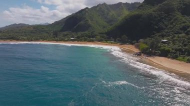 Hawaii 'de turkuaz dalgalar, altın kum, yemyeşil yemyeşil ve orman dağlarıyla dolu bir Kauai sahili. Kamera başınızın üzerinde sorunsuzca süzülüyor.