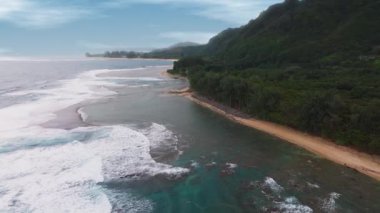Hawaii 'deki Kauai Adası' nın kıyı şeridini gösteren hava manzarası yemyeşil, engebeli kayalıklar, kumlu plajlar ve düz hareket eden okyanus dalgalarını gösteriyor..