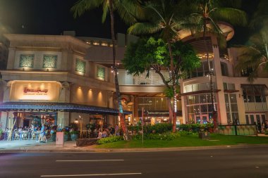 Illuminated Cheesecake Factory building in Honolulu, Hawaii, with outdoor diners, palm trees, and a well lit street in a tropical setting. clipart