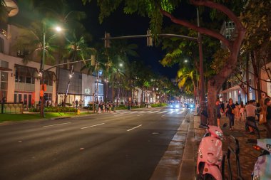 Gece Honolulu, Hawaii 'de canlı bir Waikiki caddesi. Palmiye ağaçları yol kenarında, modern binalar ev dükkanları, ve insanlar canlı bir atmosferde geziniyorlar..