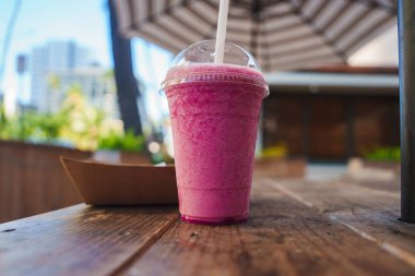 Beyaz bir pipetle berrak bir fincanda canlı pembe bir meyve püresi kırsal bir ahşap masada oturur. Bulanık yeşillik, modern bir bina ve çizgili şemsiye görülebilir..