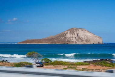 View of Rabbit Island's dome like shape off Oahu's coast, with a coastal park featuring a picnic table, parked car, and rocky shoreline. clipart