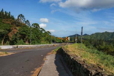 Yeşilliklerle çevrili manzaralı bir yol ve solda taş bir duvar kıvrımları, arka planda tepeler ve dağlar dağınık bulutlarla dolu berrak bir gökyüzünün altında..