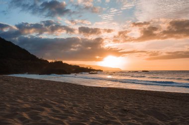 Altın ışık günbatımında okyanus dalgalarını yansıtır, kayalık çıkıntılar, yemyeşil ve uzak siluetli tepelerle kumlu bir sahili aydınlatır..