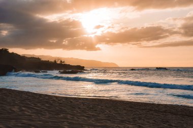 Gün batımında sakin bir Hawaii plajı. Bulutların arasından süzülen altın ışık, uzaktaki kayalık uçurumlar ve kumlu sahil boyunca hafif dalgalar..