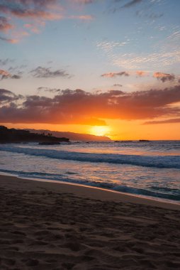 Altın turuncu günbatımı gökyüzünü ve okyanus dalgalarını aydınlatıyor, yumuşak kumlu kıyı şeridi, görünür ayak izleri ve Hawaii 'deki kayalık çıkıntılar.
