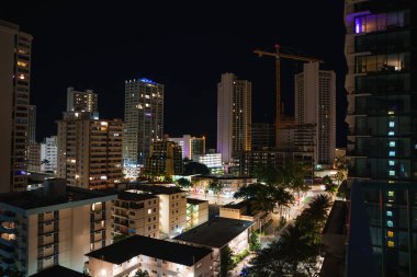 A vibrant nightscape of Honolulu, Hawaii, featuring lit high rise buildings, a construction crane, palm lined streets, and a starry sky above. clipart