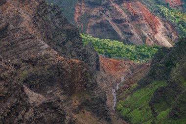 Waimea Kanyonu 'nun sarp kayalıkları yemyeşil yeşilliklerle çevrili kırmızı, kahverengi ve yeşil katmanları bir dere veya şelale ile kesiyor..