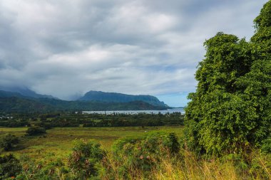 Bereketli yeşil bitki örtüsü Hawaii 'deki Kauai Adası' ndaki Napali uçurumlarını ve Hanalei Körfezi 'ni çerçeveler. Bulutlu gökyüzünün altındaki vadide küçük bir yerleşim yeri var..