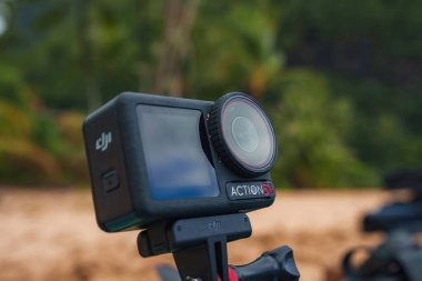 DJI Action 3 kamera dengeleyici bir kavrama üzerine monte, yemyeşil ve kumlu zemin bulanık bir zemin üzerine kurulmuş, tropik bir konum önerir.