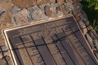 Plaque mounted on a stone structure honoring Ali i  Aimoku Kaumuali i and Moi Wahine Kekaiha akulou, surrounded by greenery in an outdoor setting. clipart