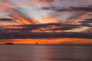 Sakin okyanus sularının üzerinde turuncu, pembe ve mor renkli canlı günbatımı. Üçgen yelkenli de dahil olmak üzere bir yolcu gemisi ve yelkenliler görülebilir..