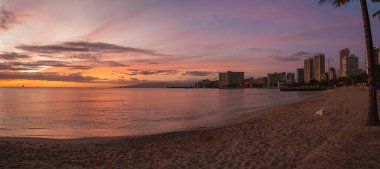Honolulu, Hawaii 'deki Waikiki Sahili üzerinde canlı bir günbatımı. Renkli gökyüzü, sakin okyanus, yelkenliler, palmiye ağaçları ve ikonik Waikiki silueti..