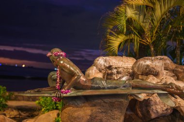Sörf tahtasındaki bir sörfçünün bronz heykeli çiçek kolyeleriyle süslenmiş kayalara, tropikal bitkilere ve karanlık gökyüzünün altındaki bir okyanus zeminine kurulmuş..