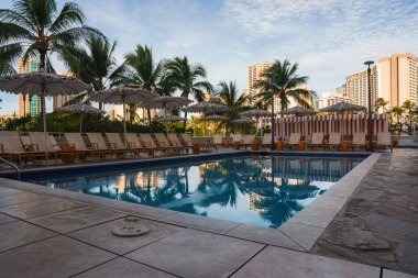 Serene havuzu şezlonglar, saman şemsiyeler ve çizgili kabinler, palmiye ağaçları ile çevrili. Arka planda görünen yüksek binalar.