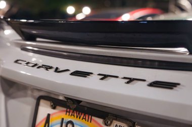 Corvette amblemini gösteren beyaz bir Corvette 'in arka görüntüsü ve yumuşak bulanık bir gece park alanında çekilmiş bir Hawaii plakası..
