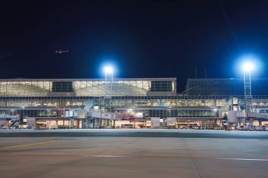 Parlak ışıkları, jet köprüleri, cam bir cephe, bir pist ve gökyüzünde küçük bir uçak olan çok katlı bir havaalanı terminalinin gece görüşü..