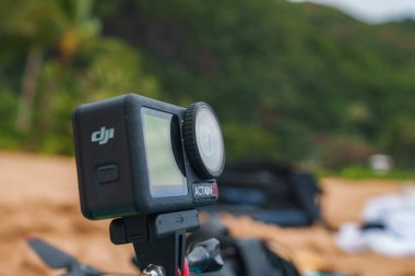 DJI Action 5 kamerası kumlu plajın bulanık arka planına ve yemyeşil bitkilere sahip bir tripoda monte edilmiştir..