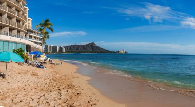 Temiz suları, renkli şemsiyeleri ve arka planda Diamond Head krateri olan altın kumsal. Ufukta bir yelkenli görünüyor..