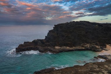 Oahu, Hawaii 'de turkuaz suları, kumlu bir plajı ve turuncudan maviye dönüşen dramatik bir gökyüzü olan karanlık volkanik kayaların kıyısı..