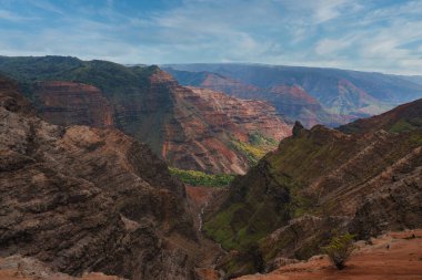 Kırmızı, yeşil ve kahverengi kayalıklar Kauai 'deki Waimea Kanyonu' na hakim. Dar bir dere, bulutlarla dolu açık bir gökyüzünün altında kanyon zemininden akar..