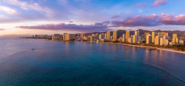 Gün batımında Honolulu, Oahu, Hawaii 'deki Waikiki Sahili' nin havadan görüntüsü. Yüksek binalar kıyı şeridi boyunca uzanır. Dağlar ve arka planda renkli gökyüzü..