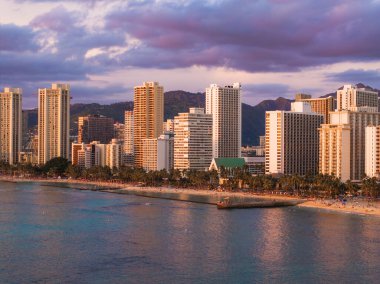Honolulu, Oahu, Hawaii 'deki Waikiki Sahili, gün batımında sakin okyanus suları, palmiye çizgili kıyılar, yüksek binalar ve uzak dağlık arazi..