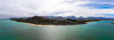 Hawaii, Oahu 'nun havadan görünüşü, kumlu bir sahil, turkuaz sular, mercan resifleri, yerleşim evleri ve bulutlu bir gökyüzünün altında yemyeşil dağlar gösteriyor..