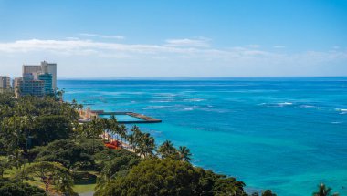 Honolulu, Oahu, Hawaii 'deki Waikiki Sahili' nin hava manzarasında turkuaz sular, küçük bir iskele, yüksek binalar ve palmiye ağaçları ile yemyeşil alanlar yer alıyor..