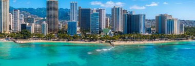Turkuaz sular Waikiki Sahili 'nde kumlu kıyılarla buluşur. Palmiye ağaçları, sahile gidenler, modern gökdelenler ve arka planda yemyeşil dağlar vardır..
