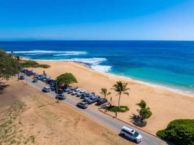 Hawaii, Oahu adasındaki bir kumsalın havadan görünüşü, altın kum, turkuaz okyanus, park etmiş arabaların, palmiye ağaçlarının ve açık gökyüzünün olduğu dar bir yol..