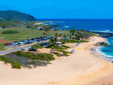 Hawaii, Oahu adasındaki kumlu bir sahil manzarası, turkuaz sular, dolambaçlı bir yol, palmiye ağaçları ve arka planda yemyeşil bir manzara..