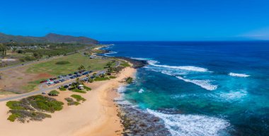 Turkuaz suları, dolambaçlı kıyı yolu, park etmiş arabalar, palmiye ağaçları ve Oahu Adası 'ndaki açık gökyüzünün altındaki yeşil tepelerin havadan görünüşü..